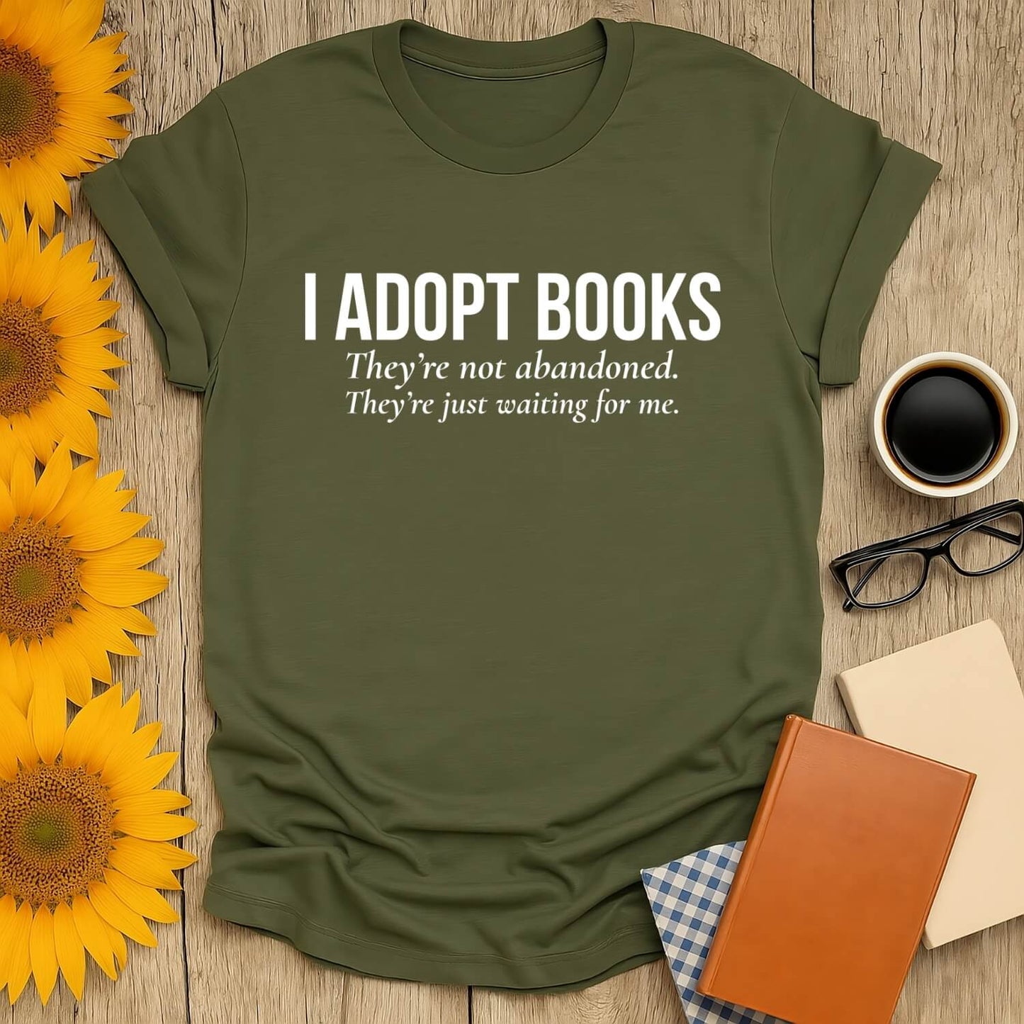 Book lover t-shirt featuring the phrase 'I adopt books' printed on a green shirt, surrounded by sunflowers and coffee.
