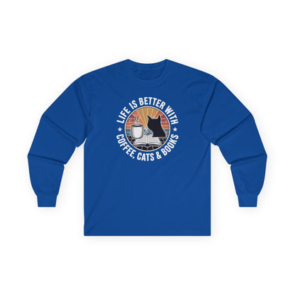 Sunset Reads & Whiskers Long Sleeve featuring cozy graphic of a cat, coffee, and books on a blue long sleeve shirt.