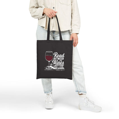 A person holding a black tote bag with the text 'Read Between the Wines' and a wine glass illustration, perfect for book lovers.