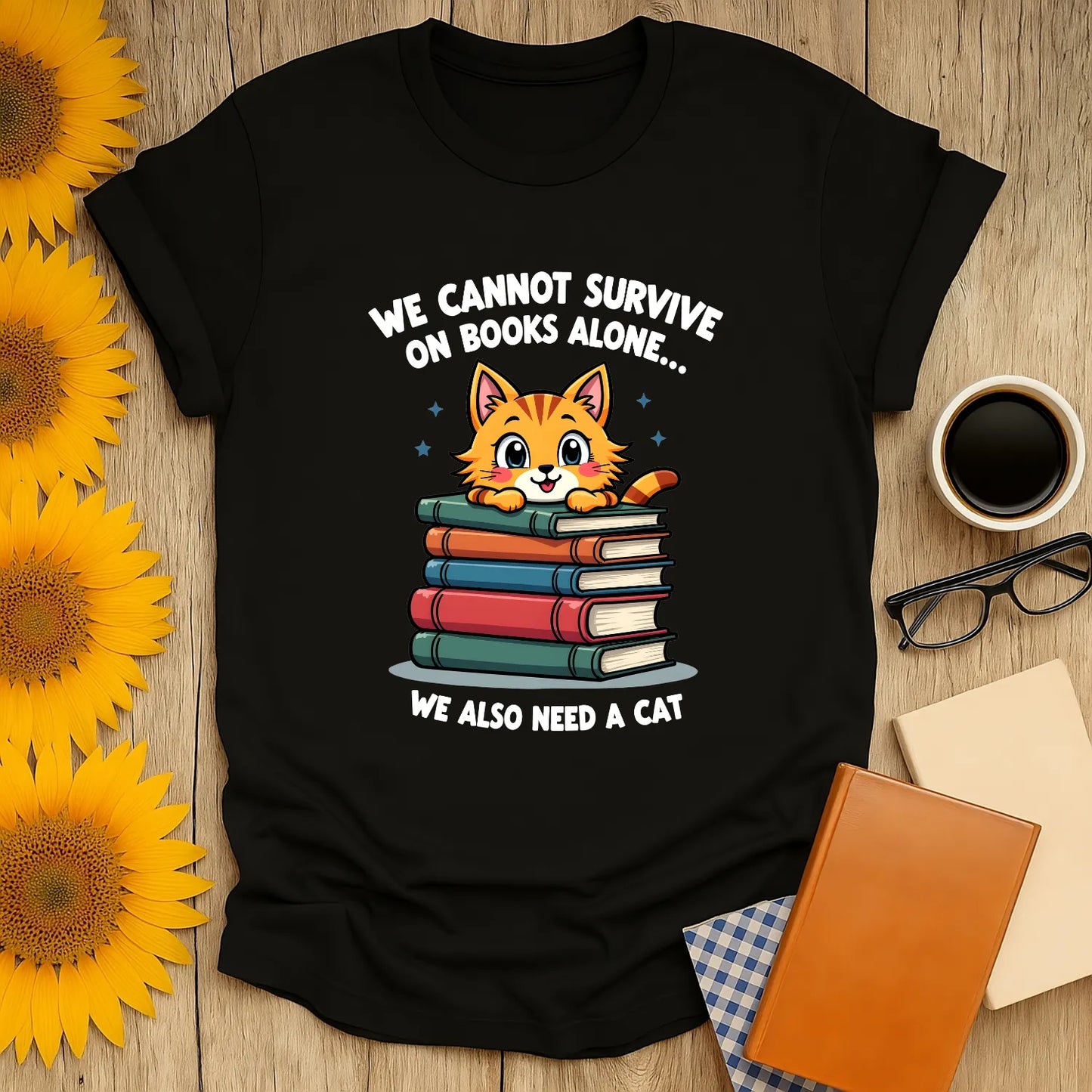 Cozy black tee featuring a cute cat atop stacked books, saying "We cannot survive on books alone... we also need a cat!" Perfect for book lovers.