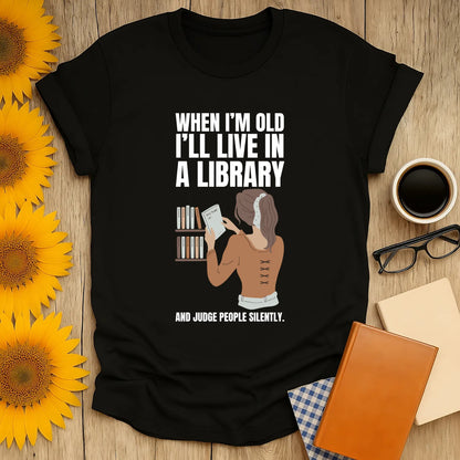 Funny book lover shirt featuring a woman reading in a library with text: 'When I'm old I'll live in a library and judge people silently.'