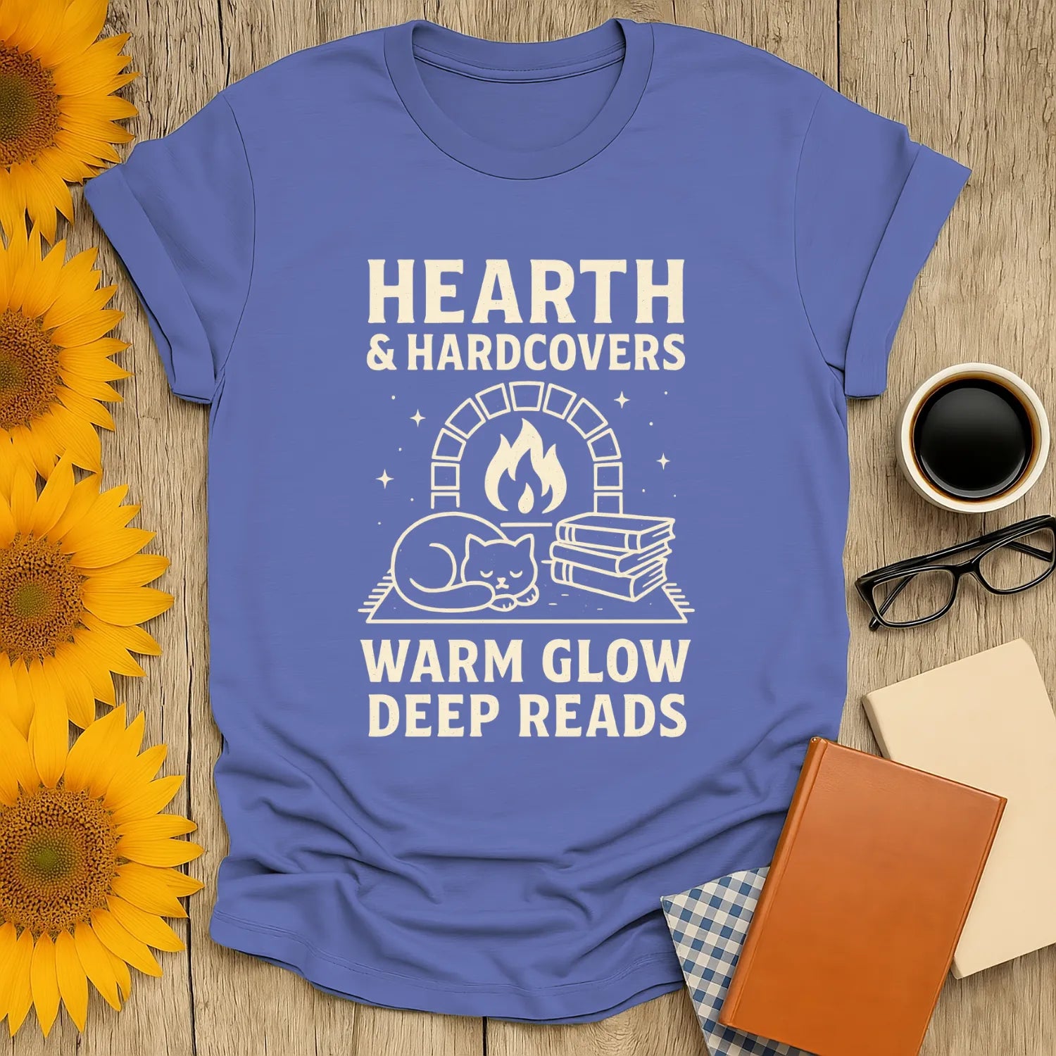 Illustrated blue tee with a cozy cat by a hearth, text: "Hearth & Hardcovers, Warm Glow Deep Reads"—perfect for book lovers!