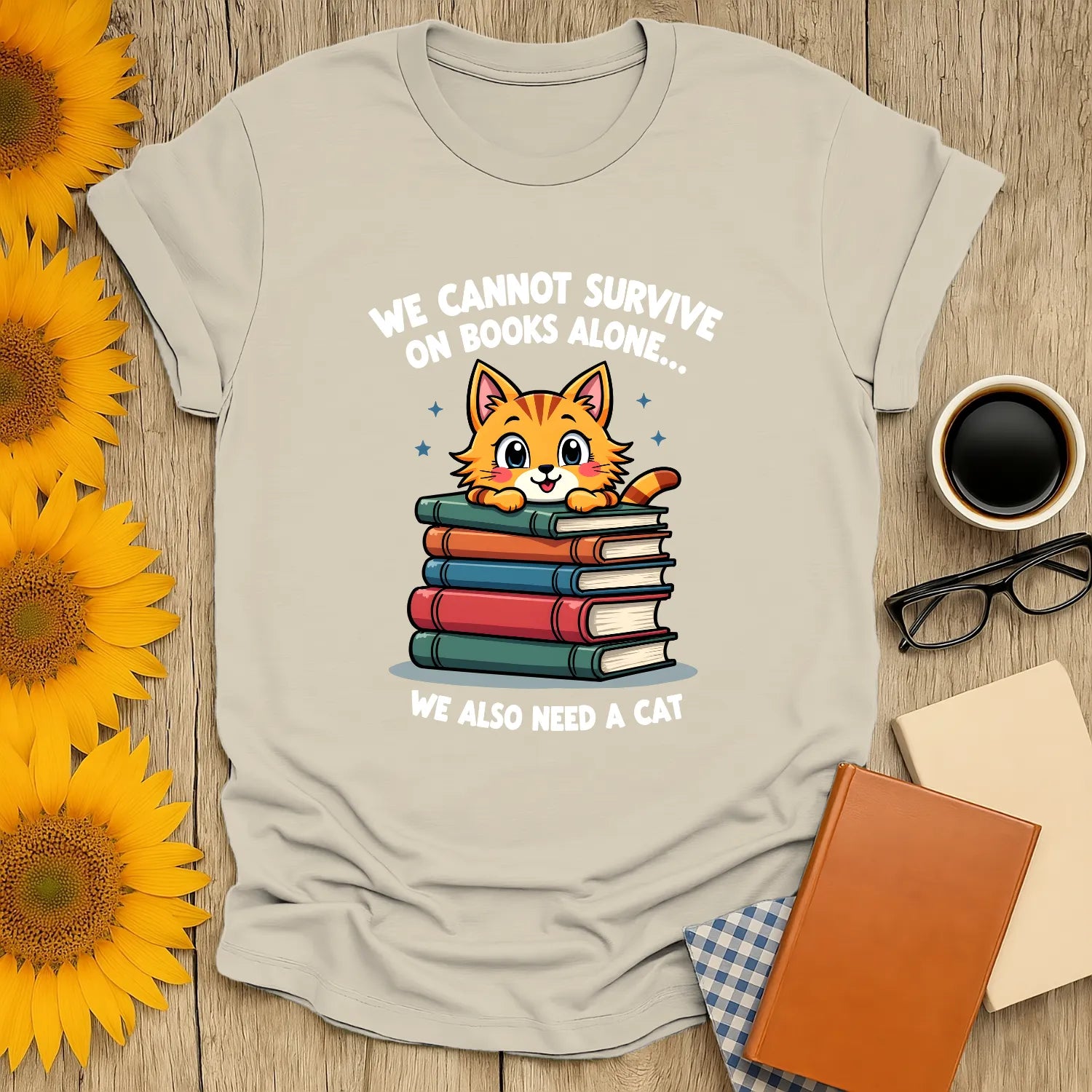 Cozy t-shirt featuring a cute cat atop stacked books with the text, 'We cannot survive on books alone… We also need a cat'.