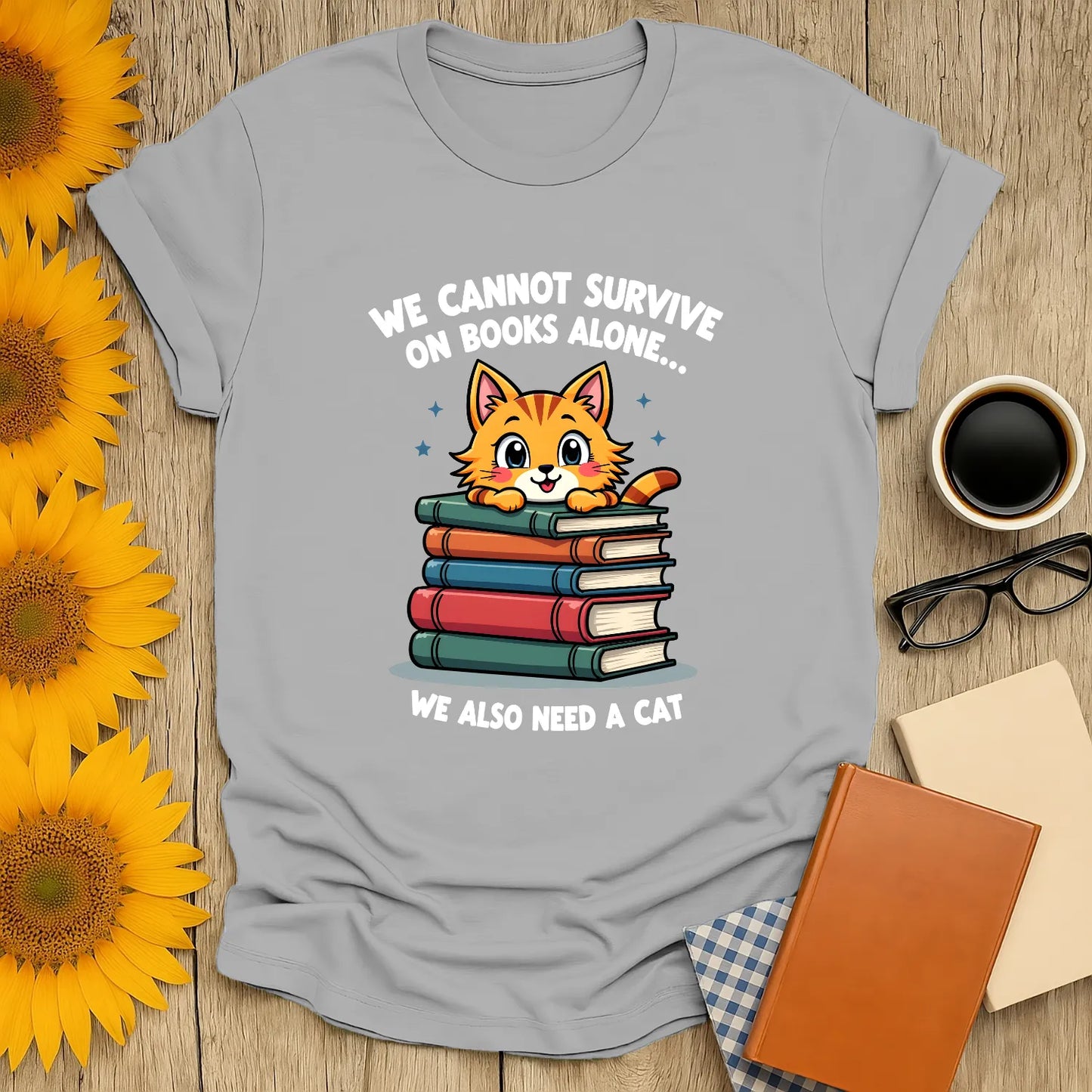 Funny reading tee featuring a cute cat atop book stacks, text: "We cannot survive on books alone... we also need a cat."