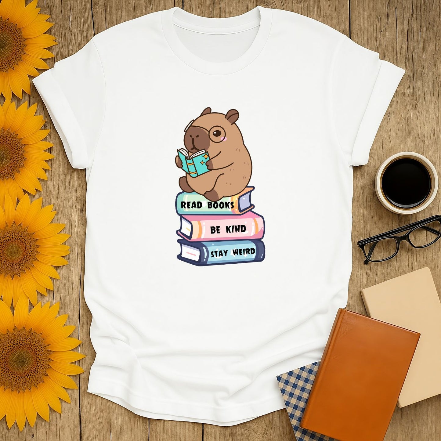 Cute capybara reading a book on stacked books with text 'Read Books, Be Kind, Stay Weird'—a cozy tee for book lovers.