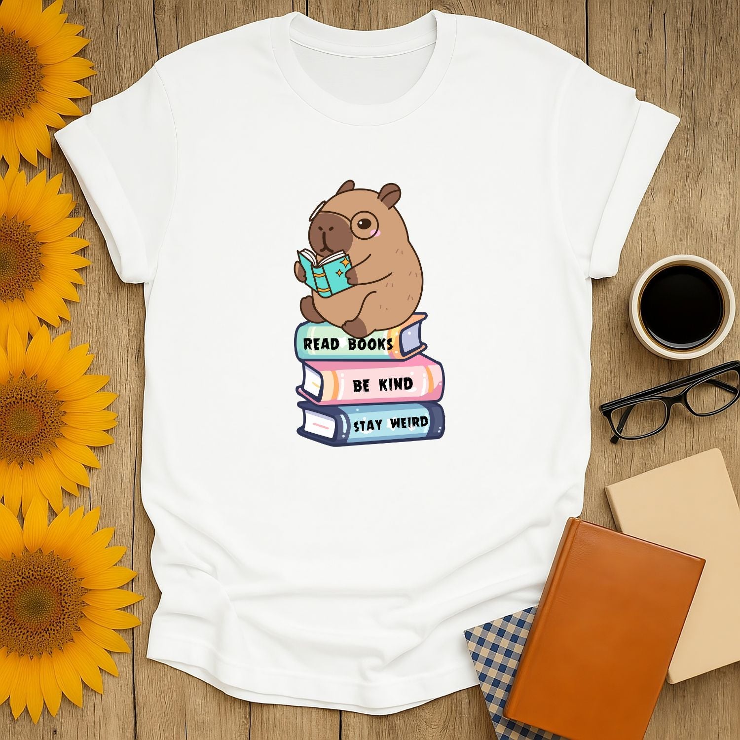 Cute capybara reading a book on stacked books with text 'Read Books, Be Kind, Stay Weird'—a cozy tee for book lovers.